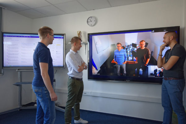 Mehrere Personen stehen vor einem Monitor während Videokonferenz im verteilten Team