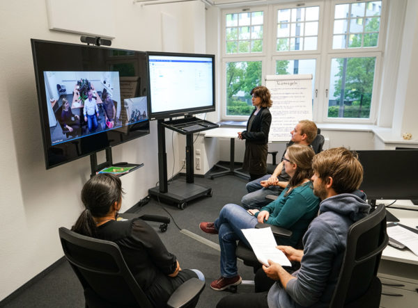 Mehrere Personen sitzen vor zwei großen Monitoren, auf denen einerseits ein weiteres Team von Personen per Videokonferenz zugeschaltet ist sowie andererseits eine Aufgabenliste zu sehen ist.