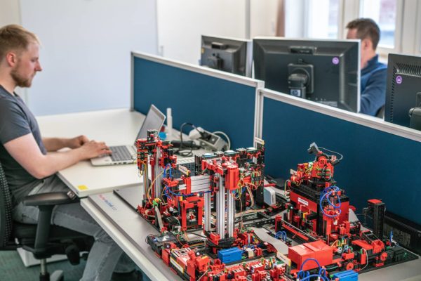 Schreibtisch auf dem eine Lernfabrik aus Lego aufgebaut ist, daneben arbeiten zwei Personen am Rechner